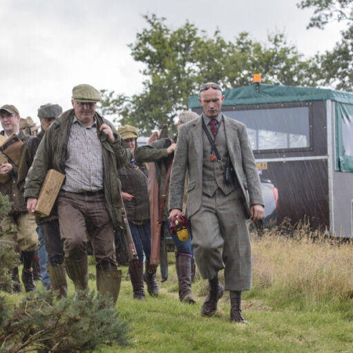 utdoor scene showing a group of people walking along a grassy path in wet weather, carrying equipment and boxes across an open countryside setting. At the front of the group, one person wears a long grey overcoat and formal-style clothing, while others behind are dressed in outdoor jackets, hats and Wellington boots. Several people are carrying rectangular wooden boxes and long items that resemble equipment cases.