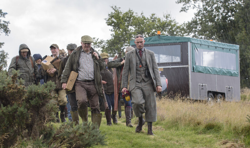 utdoor scene showing a group of people walking along a grassy path in wet weather, carrying equipment and boxes across an open countryside setting. At the front of the group, one person wears a long grey overcoat and formal-style clothing, while others behind are dressed in outdoor jackets, hats and Wellington boots. Several people are carrying rectangular wooden boxes and long items that resemble equipment cases.