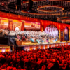 Large-scale live concert performance taking place in an ornate indoor arena before a packed audience. The foreground is filled with rows of seated spectators, illuminated by warm red and orange stage lighting. Across the front of the stage, musicians, singers and performers are arranged amongst floral decorations and festive displays.