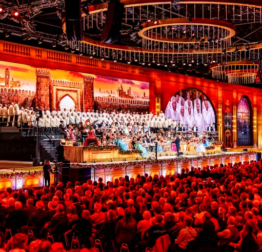 Large-scale live concert performance taking place in an ornate indoor arena before a packed audience. The foreground is filled with rows of seated spectators, illuminated by warm red and orange stage lighting. Across the front of the stage, musicians, singers and performers are arranged amongst floral decorations and festive displays.