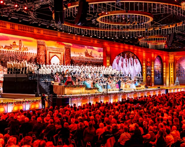 Large-scale live concert performance taking place in an ornate indoor arena before a packed audience. The foreground is filled with rows of seated spectators, illuminated by warm red and orange stage lighting. Across the front of the stage, musicians, singers and performers are arranged amongst floral decorations and festive displays.