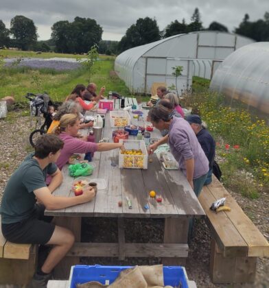 Community volunteers and growers share a meal at a large outdoor wooden picnic table beside polytunnels in a market garden setting. Crates of freshly harvested produce, drinks and lunch containers are spread across the table, while wildflowers, young trees and vegetable-growing areas surround the space. Set against open countryside with fields and mature trees under a cloudy sky, the image highlights community gardening, local food production and people coming together outdoors to enjoy a break during a day of harvesting and growing.