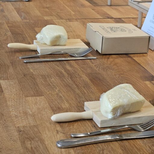 A pasta-making workshop setup is arranged on a wooden floor, with portions of dough wrapped in cling film placed on small wooden gnocchi boards beside knives and forks. Cardboard boxes and display cards for The Fife Pasta Company are positioned nearby, indicating an organised hands-on food activity. The simple layout highlights the ingredients and tools used in fresh pasta making, creating a welcoming environment for participants to learn traditional pasta-making techniques.