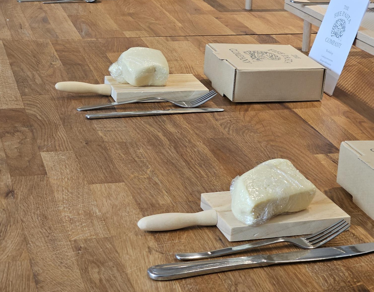 A pasta-making workshop setup is arranged on a wooden floor, with portions of dough wrapped in cling film placed on small wooden gnocchi boards beside knives and forks. Cardboard boxes and display cards for The Fife Pasta Company are positioned nearby, indicating an organised hands-on food activity. The simple layout highlights the ingredients and tools used in fresh pasta making, creating a welcoming environment for participants to learn traditional pasta-making techniques.