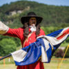 Historical re-enactment scene featuring a costumed interpreter dressed in a bright red period coat with large cream cuffs, a wide-brimmed black hat and a long curled wig, standing outdoors while holding a large Union flag. The person gestures with one arm outstretched as if addressing an audience during a living history demonstration. The setting is an open grassy field bordered by trees and wooded hills under bright sunshine, with a few spectators visible in the background. The colourful costume, historic flag and rural backdrop create the atmosphere of a heritage event celebrating local history and historical re-enactment.