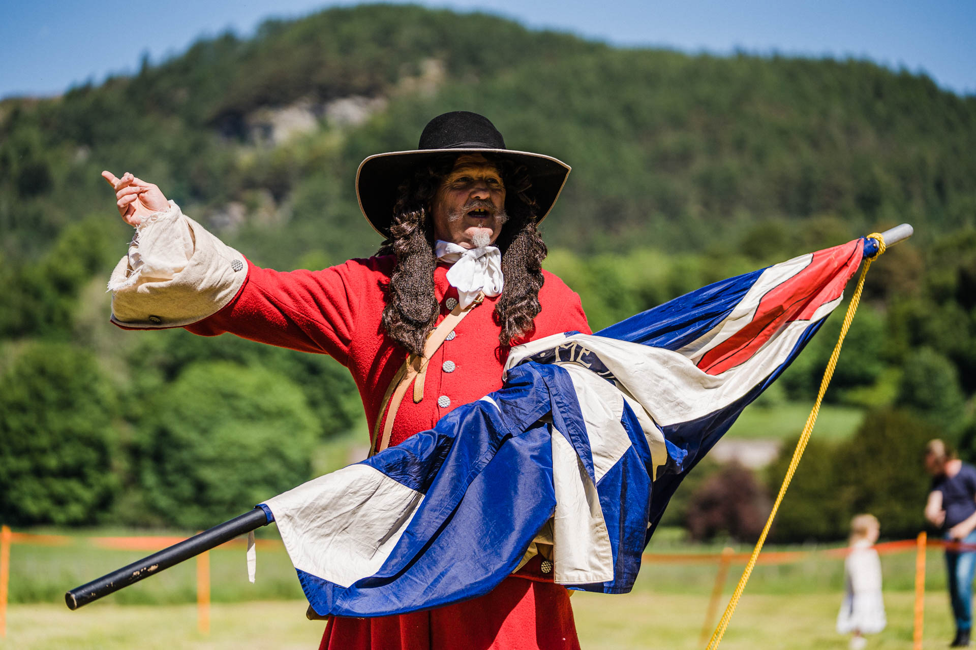 Historical re-enactment scene featuring a costumed interpreter dressed in a bright red period coat with large cream cuffs, a wide-brimmed black hat and a long curled wig, standing outdoors while holding a large Union flag. The person gestures with one arm outstretched as if addressing an audience during a living history demonstration. The setting is an open grassy field bordered by trees and wooded hills under bright sunshine, with a few spectators visible in the background. The colourful costume, historic flag and rural backdrop create the atmosphere of a heritage event celebrating local history and historical re-enactment.
