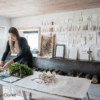 Person arranging natural materials on a worktable in a bright studio space, surrounded by botanical samples, handmade artworks and textured paper studies displayed on the wall. A row of logs sits beneath a fireplace, and woven sculptures hang near a large window. Photo credit: Sim Canetty-Clarke.