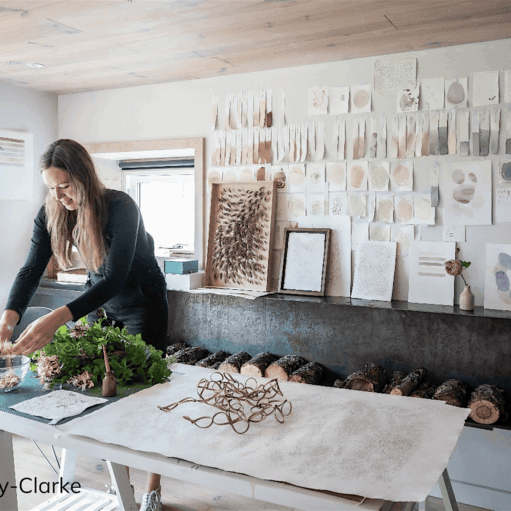 Person arranging natural materials on a worktable in a bright studio space, surrounded by botanical samples, handmade artworks and textured paper studies displayed on the wall. A row of logs sits beneath a fireplace, and woven sculptures hang near a large window. Photo credit: Sim Canetty-Clarke.