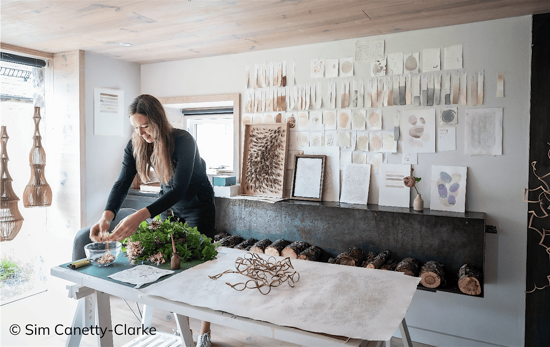 Person arranging natural materials on a worktable in a bright studio space, surrounded by botanical samples, handmade artworks and textured paper studies displayed on the wall. A row of logs sits beneath a fireplace, and woven sculptures hang near a large window. Photo credit: Sim Canetty-Clarke.