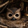 A detailed wooden owl mask with large, hollow eye cutouts and a pointed black beak, resting on fur and surrounded by natural forest elements.