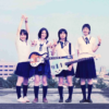 Four high school girls in matching Japanese sailor-style uniforms posing together on an open rooftop, with two holding electric guitars and flashing peace signs.