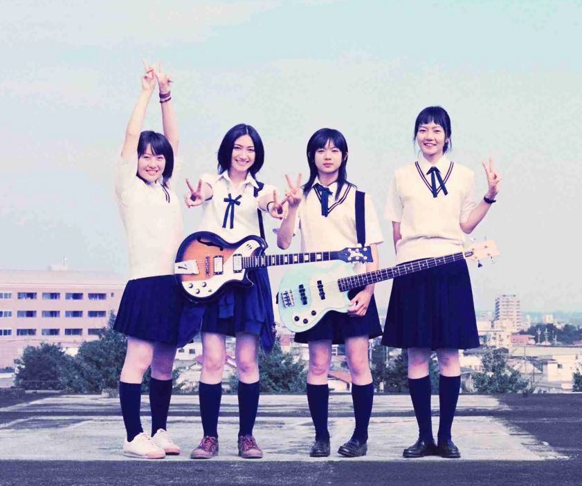 Four high school girls in matching Japanese sailor-style uniforms posing together on an open rooftop, with two holding electric guitars and flashing peace signs.