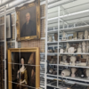 A storage room or archive containing art collections, with framed portraits hanging on mesh sliding racks on the left and various sculptures, busts, and artifacts arranged on white shelving units on the right.