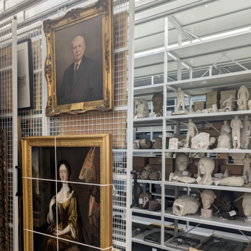 A storage room or archive containing art collections, with framed portraits hanging on mesh sliding racks on the left and various sculptures, busts, and artifacts arranged on white shelving units on the right.