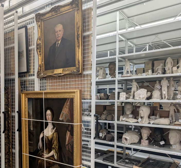 A storage room or archive containing art collections, with framed portraits hanging on mesh sliding racks on the left and various sculptures, busts, and artifacts arranged on white shelving units on the right.
