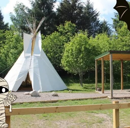An outdoor area featuring a large white teepee tent set against a background of lush green trees, decorated with cartoon Halloween graphics including a mummy in the bottom left corner and a bat in the top right.