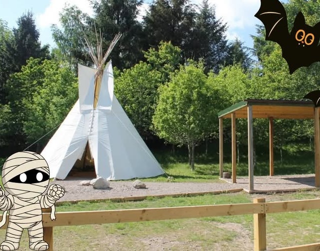 An outdoor area featuring a large white teepee tent set against a background of lush green trees, decorated with cartoon Halloween graphics including a mummy in the bottom left corner and a bat in the top right.