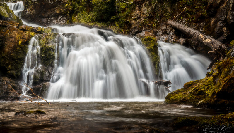 A long-exposure photograph of cascading white waterfalls flowing over dark, mossy rocks in a forested gorge.