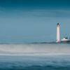 A tall, white lighthouse standing on a distant coast, viewed across silky, long-exposure ocean waves under a blue sky.