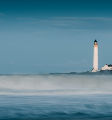 A tall, white lighthouse standing on a distant coast, viewed across silky, long-exposure ocean waves under a blue sky.