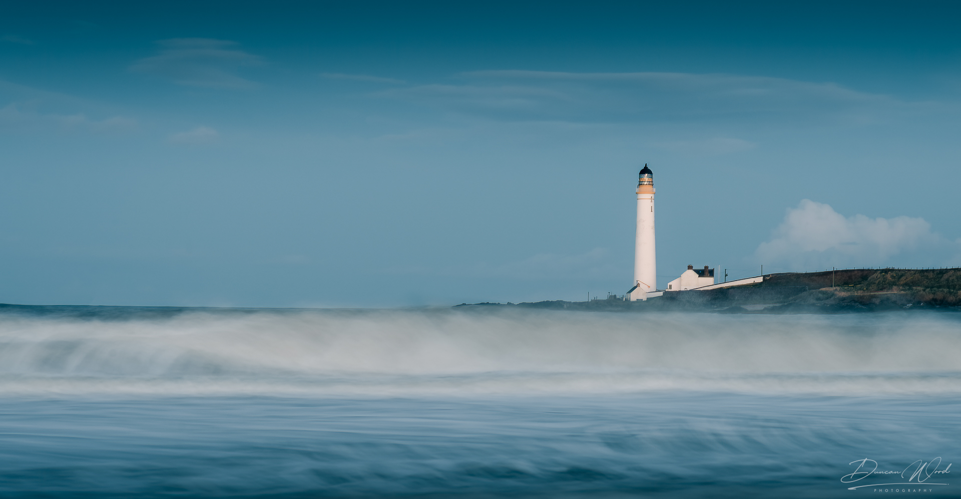 A tall, white lighthouse standing on a distant coast, viewed across silky, long-exposure ocean waves under a blue sky.