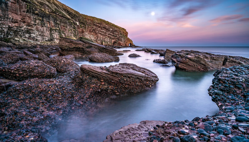 A rocky, pebbled beach with large stone formations in mist-like water, set against steep coastal cliffs under a twilight sky with a bright moon.