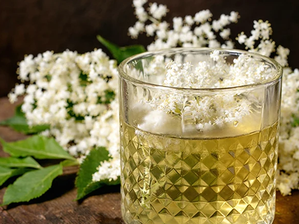 A textured glass filled with light yellow elderflower drink and ice cubes, garnished with fresh elderflower blossoms, resting on a rustic wooden surface next to elderflower sprigs and green leaves.