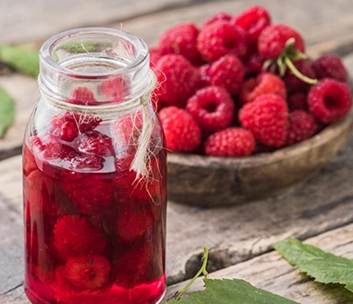 A glass jar tied with twine filled with red raspberry liquid and submerged berries, sitting on a rustic wooden table next to a wooden bowl piled high with fresh raspberries.