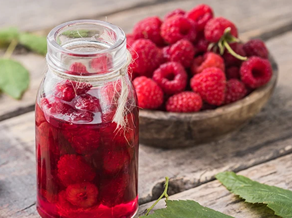 A glass jar tied with twine filled with red raspberry liquid and submerged berries, sitting on a rustic wooden table next to a wooden bowl piled high with fresh raspberries.