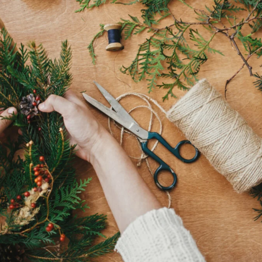 Hands crafting a holiday wreath on a wooden surface with pine branches, red berries, scissors, and a roll of twine.