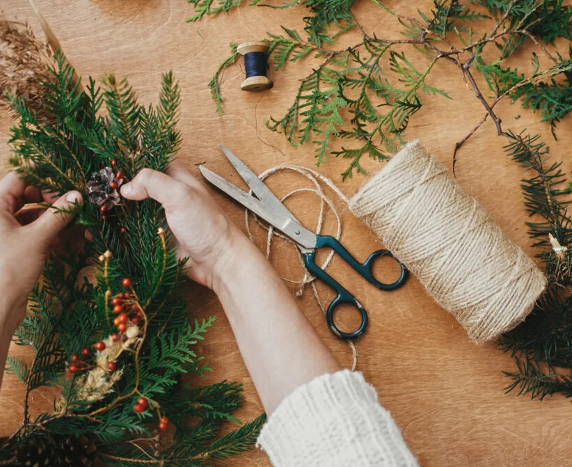 Hands crafting a holiday wreath on a wooden surface with pine branches, red berries, scissors, and a roll of twine.
