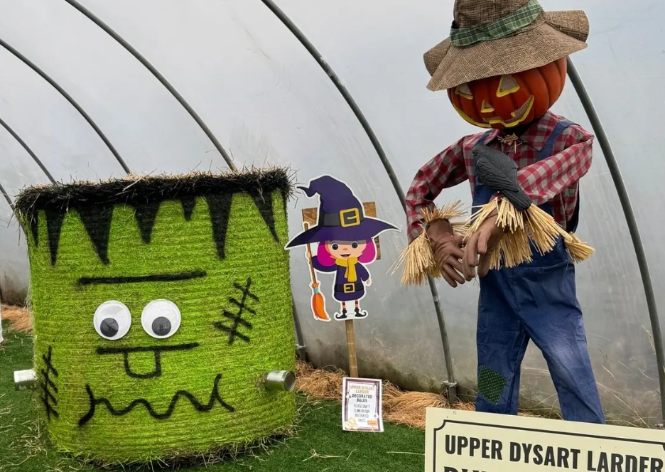 A Halloween display featuring a green, hay-bale Frankenstein's monster with googly eyes alongside a pumpkin-headed scarecrow in overalls and a straw hat inside a polytunnel greenhouse.