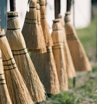 A row of handmade straw brooms tied with black twine, shown hanging outdoors in soft, natural light.