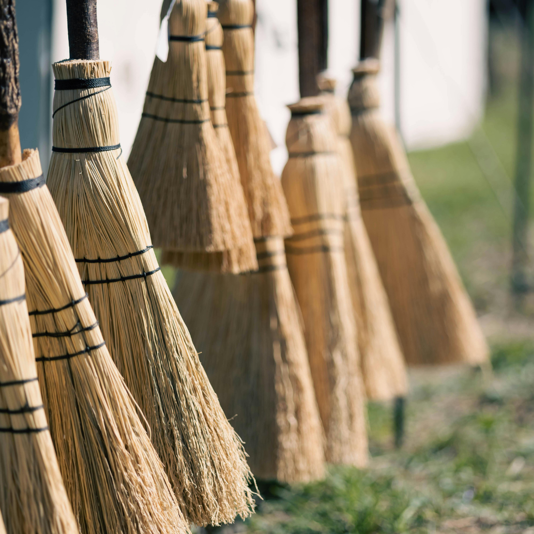 A row of handmade straw brooms tied with black twine, shown hanging outdoors in soft, natural light.
