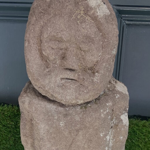 An ancient, carved stone statue depicting a human figure with simplified facial features, standing on green artificial turf against a dark grey wall background.