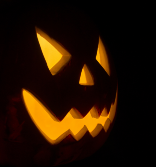 A carved jack-o'-lantern illuminated from within by a warm, yellow glow, showing a menacing face with angled eyes and a jagged smile against a pitch-black background.