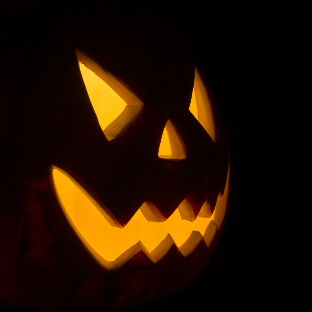 A carved jack-o'-lantern illuminated from within by a warm, yellow glow, showing a menacing face with angled eyes and a jagged smile against a pitch-black background.