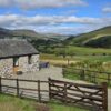 Traditional stone cottage set against green pastures and rolling hills, surrounded by a wooden post-and-rail fence and gravel driveway.