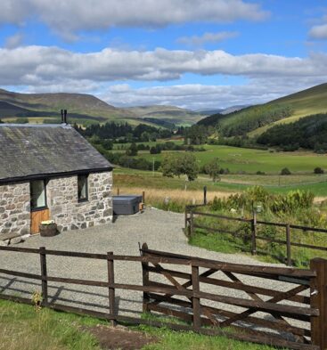Traditional stone cottage set against green pastures and rolling hills, surrounded by a wooden post-and-rail fence and gravel driveway.