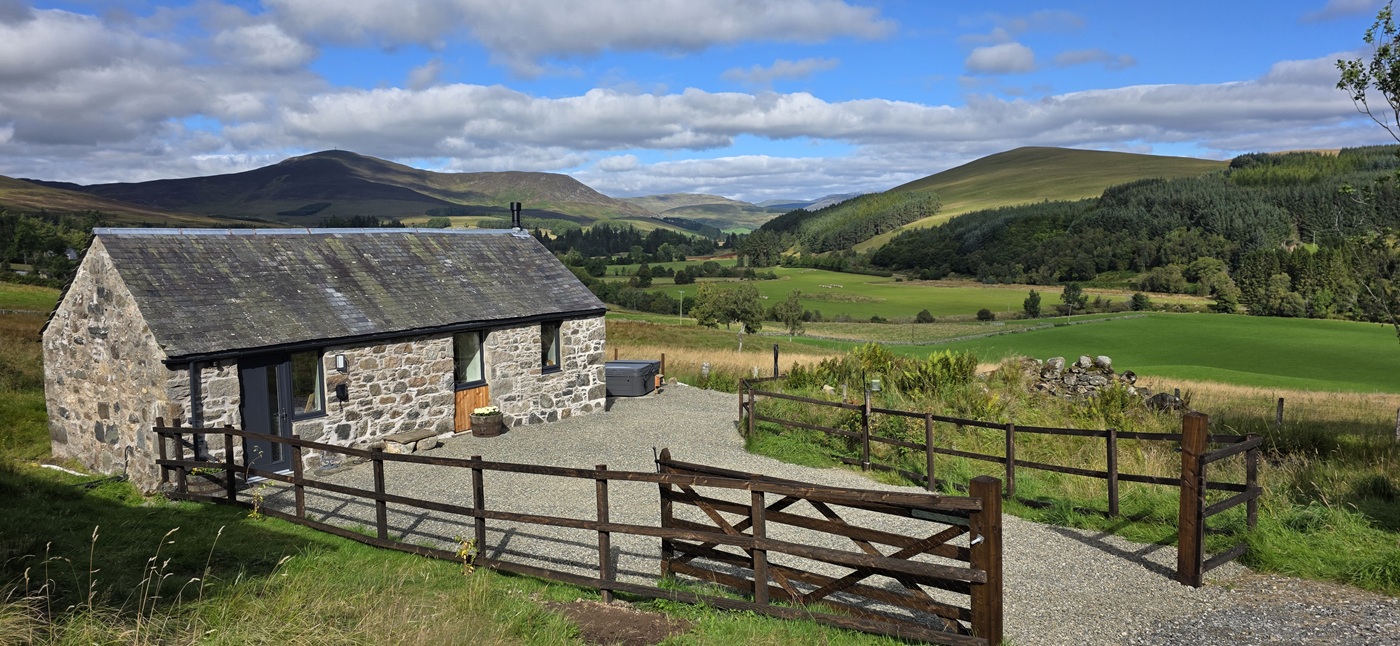 Traditional stone cottage set against green pastures and rolling hills, surrounded by a wooden post-and-rail fence and gravel driveway.