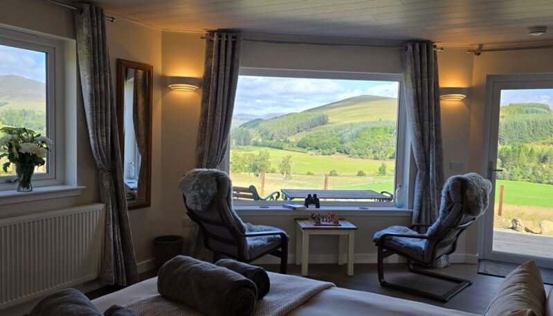 Cosy room interior featuring warm wall lights, two armchair seating spots by a large picture window, and wide views of rolling green hills.