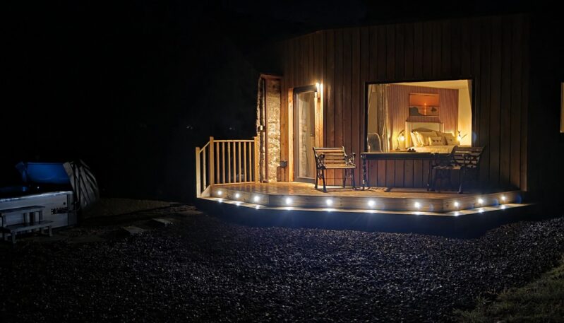 A modern wooden cabin at night featuring a deck illuminated by inset floor lights, an outdoor hot tub glowing blue to the left, and a large window showing a warmly lit bedroom interior.
