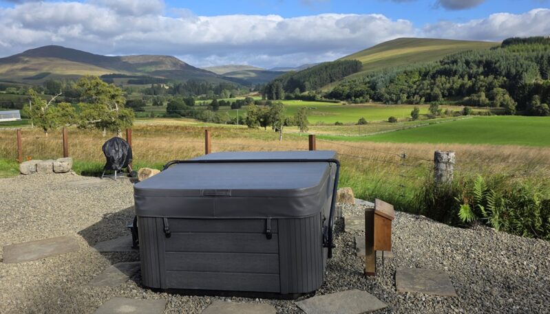 A grey covered hot tub sitting on a gravel patio next to a barbecue, with a scenic backdrop of sprawling green pastures, trees, and rolling hills under a blue sky.