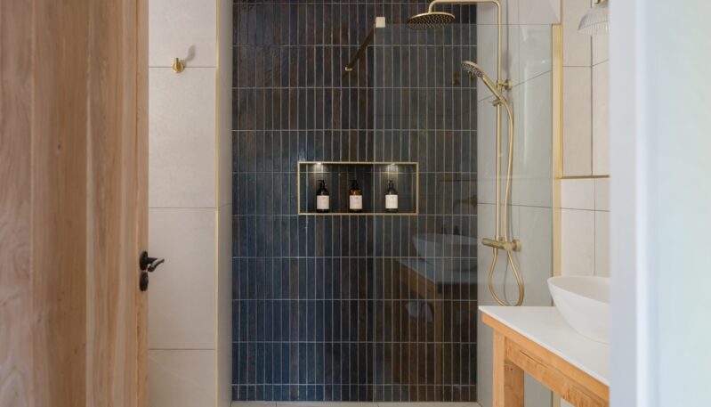Modern bathroom featuring a walk-in shower with vertical dark blue subway tiles, gold fixtures, a recessed shelf with toiletries, and a wooden vanity unit.