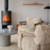 Cozy living room space with two plush, cream shearling armchairs, a smooth grey coffee table, and a modern black wood-burning stove lit in the background.