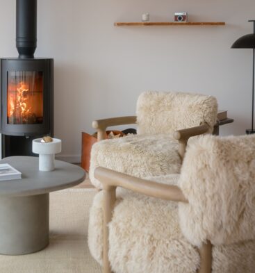 Cozy living room space with two plush, cream shearling armchairs, a smooth grey coffee table, and a modern black wood-burning stove lit in the background.
