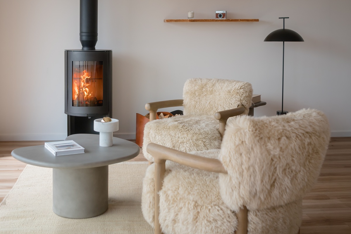 Cozy living room space with two plush, cream shearling armchairs, a smooth grey coffee table, and a modern black wood-burning stove lit in the background.