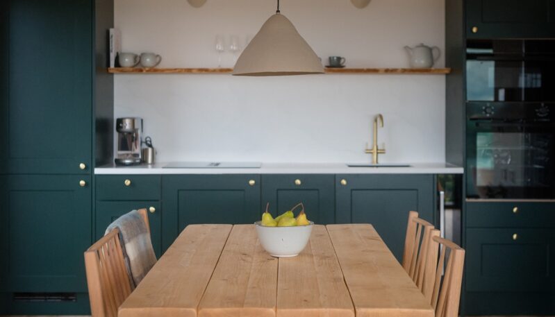 Wooden dining table set with a bowl of pears, positioned in front of a stylish dark green kitchen with brass hardware, open wooden shelving, and a clay pendant light overhead.