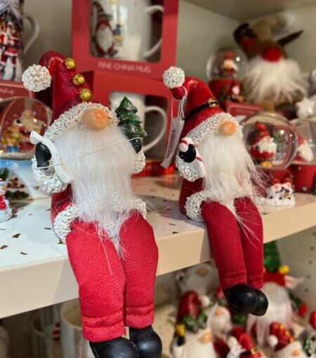 Christmas-themed shop display featuring two red-and-white Santa gnome decorations with dangling legs, surrounded by festive mugs, ornaments and seasonal gift items on shelves.