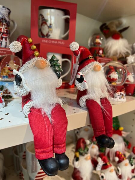 Christmas-themed shop display featuring two red-and-white Santa gnome decorations with dangling legs, surrounded by festive mugs, ornaments and seasonal gift items on shelves.